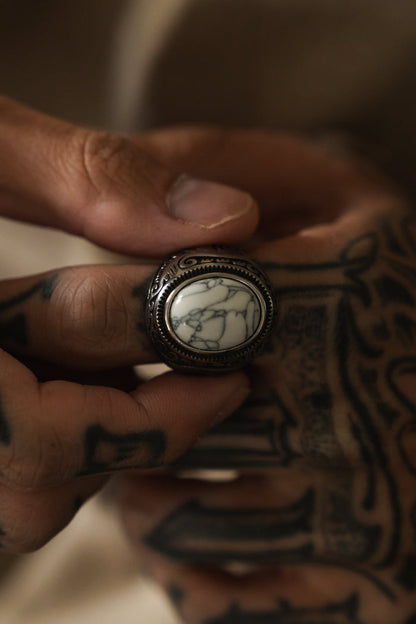 A close-up shows tattooed hands holding a Relentless Betrayal Tempest Ring. The ornate band cradles a large, smooth white stone veined with dark lines, subtly resting between thumb and forefinger, evoking elemental forces from mythical tales.