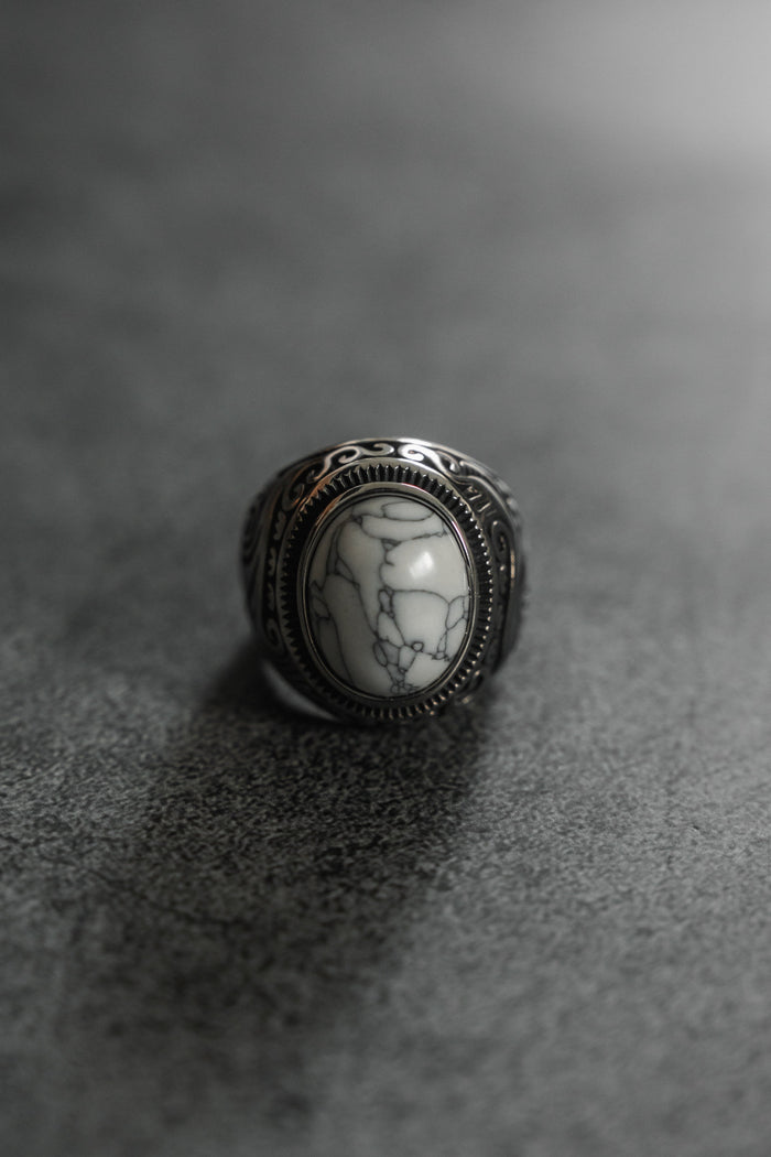The Relentless Betrayal Tempest Ring is a silver masterpiece with an intricate pattern, showcasing a large oval white stone veined with darkness. Set on a textured gray surface, its reminiscent of mythical tales and elemental forces, highlighted by lighting that accentuates its detailed design.