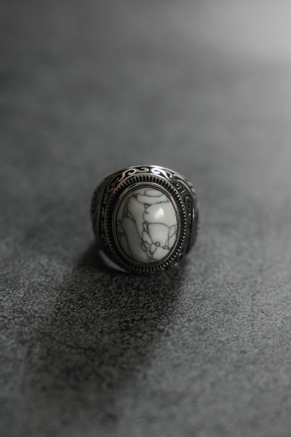 The Relentless Betrayal Tempest Ring is a silver masterpiece with an intricate pattern, showcasing a large oval white stone veined with darkness. Set on a textured gray surface, its reminiscent of mythical tales and elemental forces, highlighted by lighting that accentuates its detailed design.