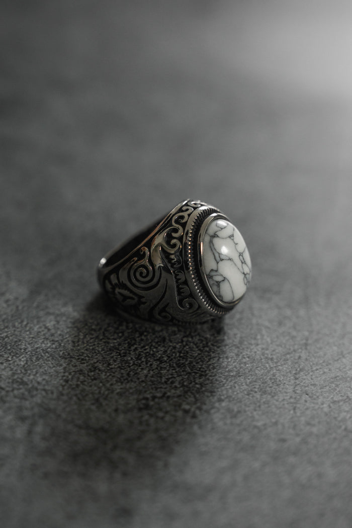 The Tempest Ring by Relentless Betrayal is a silver masterpiece featuring an intricate design and a large, oval white stone with gray marbling. Its polished finish stands out on a dark, textured background, evoking mythical themes through its exquisite craftsmanship.
