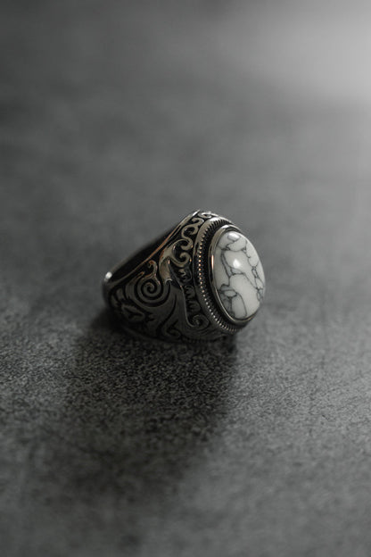 The Tempest Ring by Relentless Betrayal is a silver masterpiece featuring an intricate design and a large, oval white stone with gray marbling. Its polished finish stands out on a dark, textured background, evoking mythical themes through its exquisite craftsmanship.