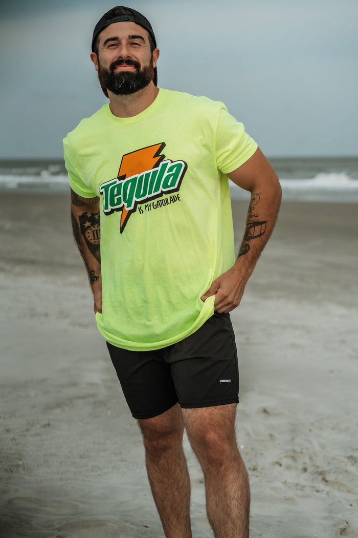 A bearded man in a neon yellow Tequila Is My Gatorade - Lemon Lime Tango Charlie Apparel t-shirt, black shorts, and a backward cap smiles on a sunny beach with the ocean behind him, showing off one of our bold best sellers.