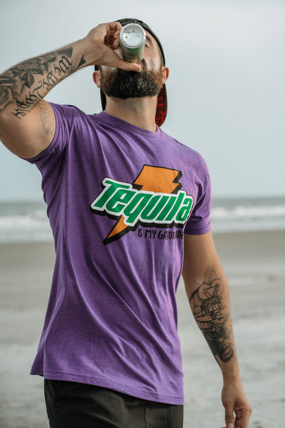 A tattooed man drinks from a bottle on the beach, wearing Tango Charlie Apparel’s “Tequila Is My Gatorade - Purple Rush” T-shirt as the ocean and sky provide a vivid backdrop.