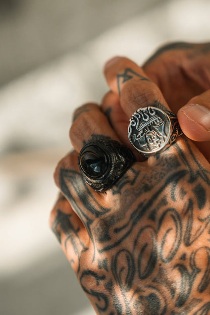 A close-up shows a tattooed hand wearing two ornate rings. One is Relentless Betrayals The Black Pearl Ring with a dark stone, and the other features an engraved face. Soft lighting highlights the detailed tattoos and intricate ring carvings.