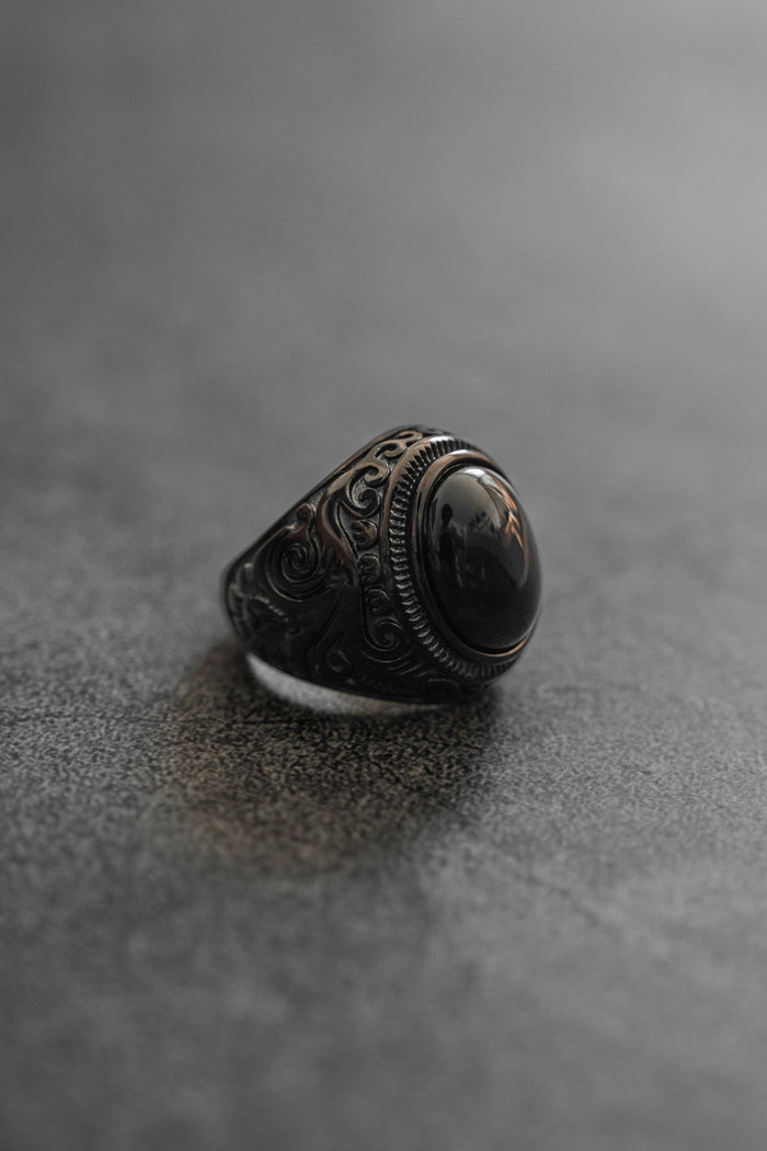 Relentless Betrayals Black Pearl Ring, featuring intricate swirling patterns and a large oval black stone, elegantly rests on a textured gray surface with a softly blurred background that highlights the stainless steel jewelrys ornate details.