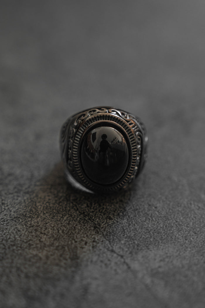 A detailed view of Relentless Betrayals The Black Pearl Ring displays a dark, intricate design featuring an oval black stone in a complex silver band, inspired by French Polynesia pearls, on a textured dark gray surface. The stone’s reflection captures a faint silhouette.