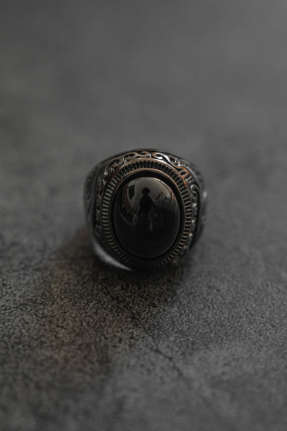 A detailed view of Relentless Betrayals The Black Pearl Ring displays a dark, intricate design featuring an oval black stone in a complex silver band, inspired by French Polynesia pearls, on a textured dark gray surface. The stone’s reflection captures a faint silhouette.