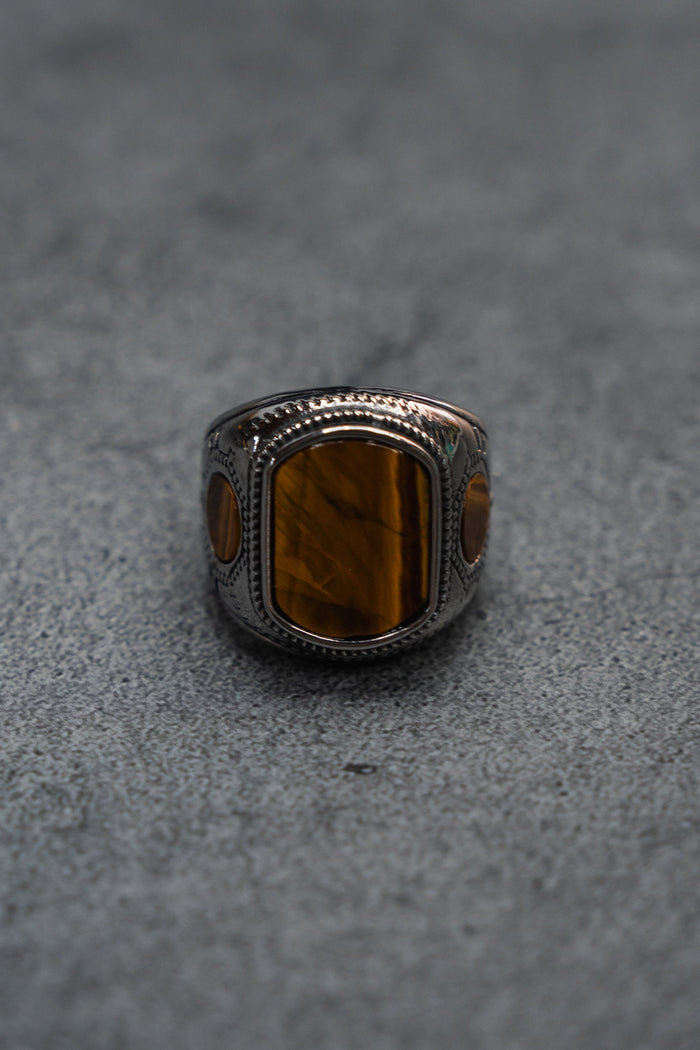 The Desert Relic Ring by Relentless Betrayal is a handcrafted silver band showcasing a rectangular brown stone at its center, beautifully presented on a minimalist gray surface.