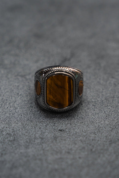 The Desert Relic Ring by Relentless Betrayal is a handcrafted silver band showcasing a rectangular brown stone at its center, beautifully presented on a minimalist gray surface.