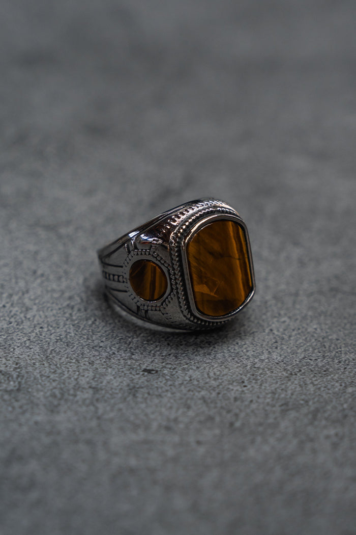 A handcrafted ring from Relentless Betrayal, The Desert Relic Ring, featuring a silver engraved band and a rectangular amber-colored stone, rests on a gray textured surface, capturing its timeless allure.