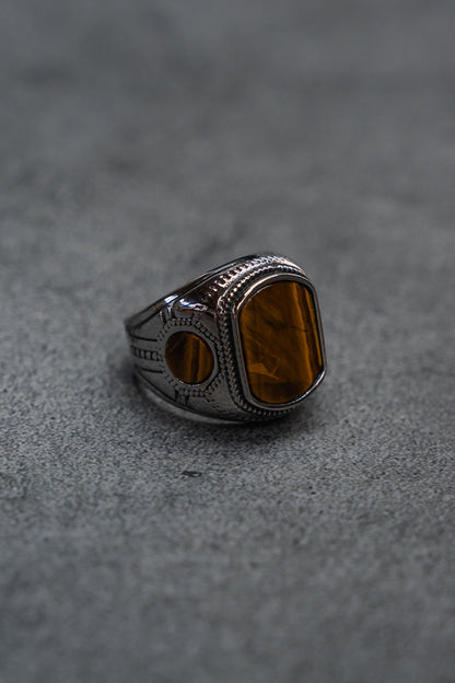A handcrafted ring from Relentless Betrayal, The Desert Relic Ring, featuring a silver engraved band and a rectangular amber-colored stone, rests on a gray textured surface, capturing its timeless allure.