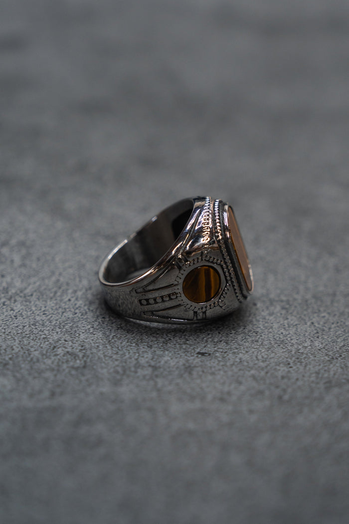 The Desert Relic Ring by Relentless Betrayal, a silver handcrafted piece featuring an oval amber-colored stone, is displayed upright on a gray textured surface and photographed from the side.