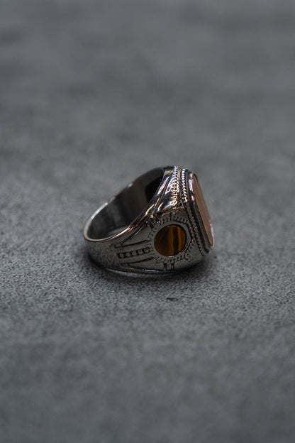 The Desert Relic Ring by Relentless Betrayal, a silver handcrafted piece featuring an oval amber-colored stone, is displayed upright on a gray textured surface and photographed from the side.