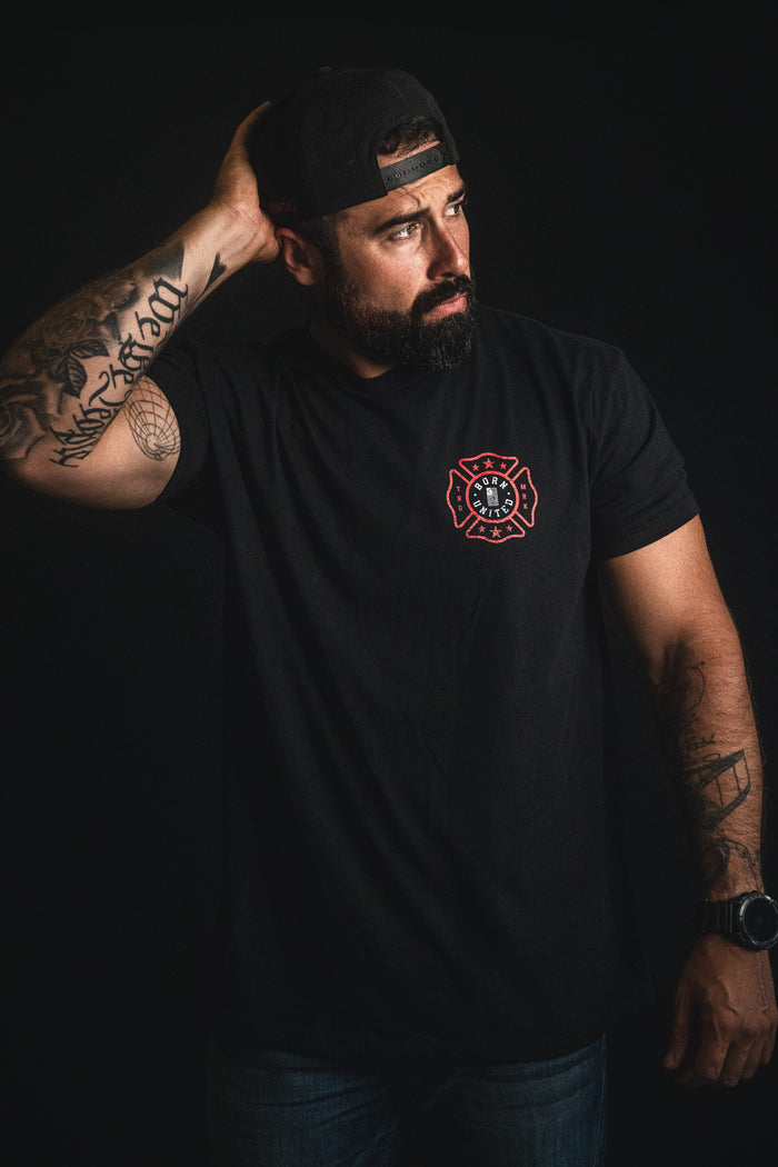 A man stands against a dark background, wearing a black t-shirt featuring The Fireman's Prayer by Born United Private Label and a backward cap, his tattooed arm resting behind his head. This powerful image makes an ideal firefighter gift or inspiration for a heartfelt firefighter poem.