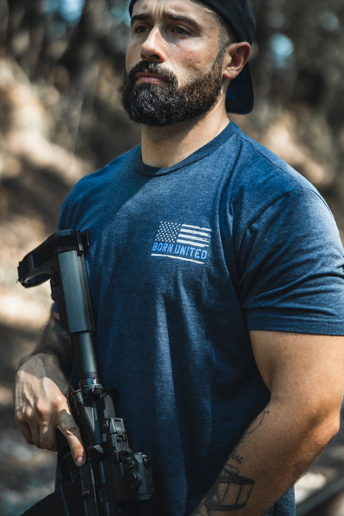 A bearded man in a backwards black cap and a blue The Good Are Unafraid T-shirt by Born United Private Label stands outside, confidently holding a rifle amid trees and sunlight—reflecting bold style and strength.