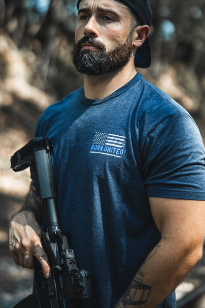 A bearded man in a backwards black cap and a blue The Good Are Unafraid T-shirt by Born United Private Label stands outside, confidently holding a rifle amid trees and sunlight—reflecting bold style and strength.