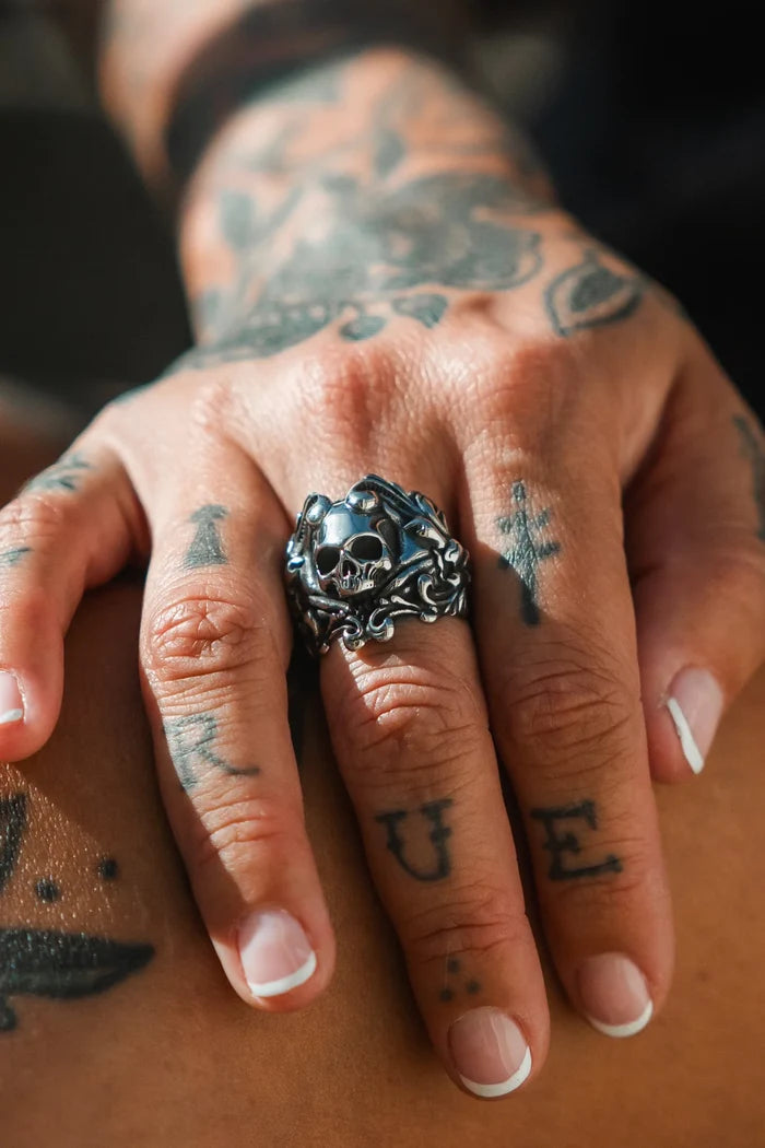 A hand with tattoos, featuring crosses and the letters L U E, wears The Gravewater Ring by Relentless Betrayal—a large, handcrafted men's skull ring—on the middle finger. The person has a French manicure.