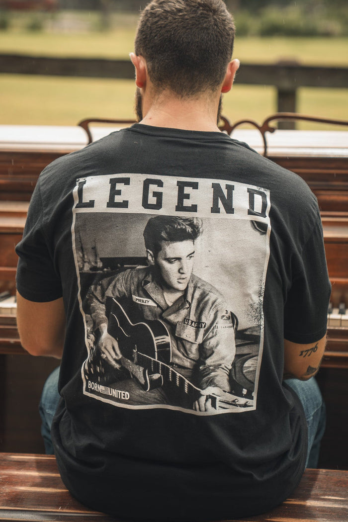 A man sits outdoors at a piano, seen from behind, wearing the Born United Private Label The King Legend Tee—a black shirt featuring a black-and-white image of Elvis Presley with LEGEND above, honoring the rock and roll icon.