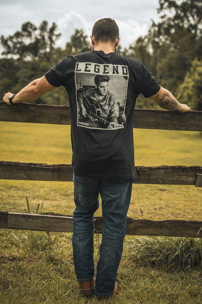 A man outdoors leans on a wooden fence, wearing the Born United Private Label The King Legend Tee, featuring a photo of a rock musician and LEGEND above it. Trees and grass are in the background.
