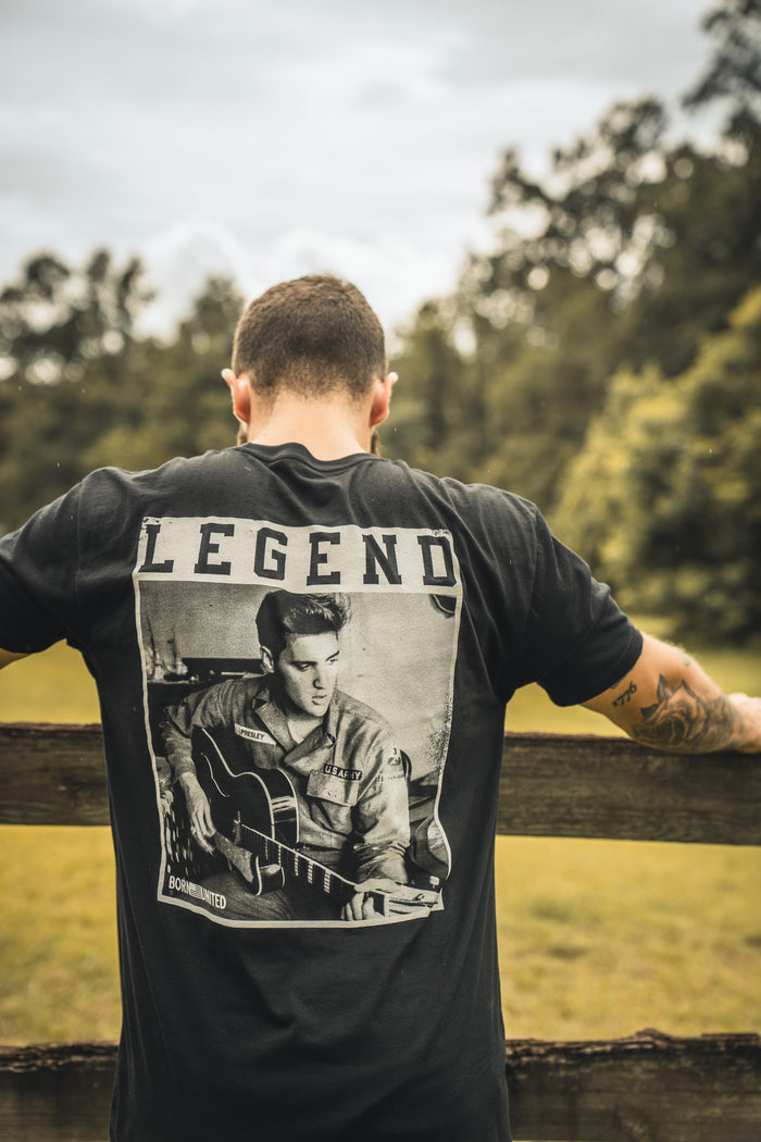 A man with short hair stands outdoors by a wooden fence, wearing the Born United Private Label The King Legend Tee featuring a guitarist and the word LEGEND. Trees and grass fill the background, celebrating rock music style.