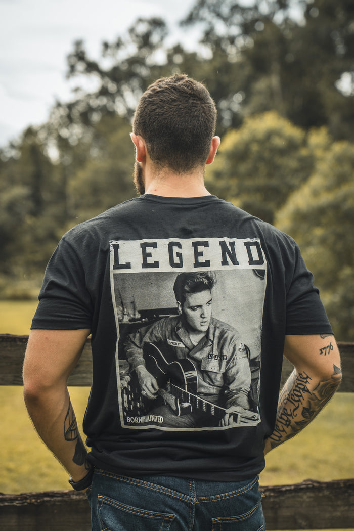 A tattooed man stands outdoors, back to the camera, wearing the Born United Private Label The King Legend Tee, a black shirt with a black-and-white photo of a guitarist and LEGEND above—a perfect tribute to rock and roll.