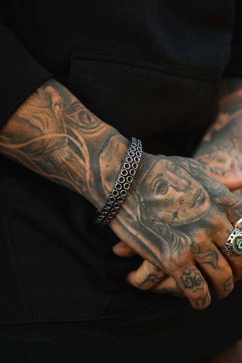 Tattooed hands are clasped, one showcasing a womans face and the other with smaller designs. The wrists feature The Kraken Bracelet by Relentless Betrayal, while multiple rings adorn the fingers. The person is dressed in a dark top.