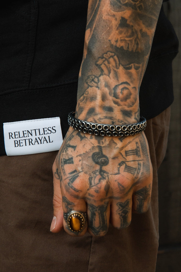 A tattooed hand showcases The Kraken Bracelet and an amber ring, paired with a vintage watch design. With a Relentless Betrayal patched black sleeve and brown pants, the look exudes rugged sophistication.
