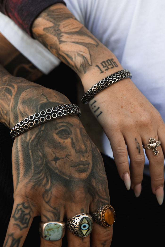 Two tattooed hands showcase Relentless Betrayals hand-forged stainless steel Kraken Bracelet on the right, alongside rings and portrait tattoos on the left. Both rest elegantly on a dark surface.