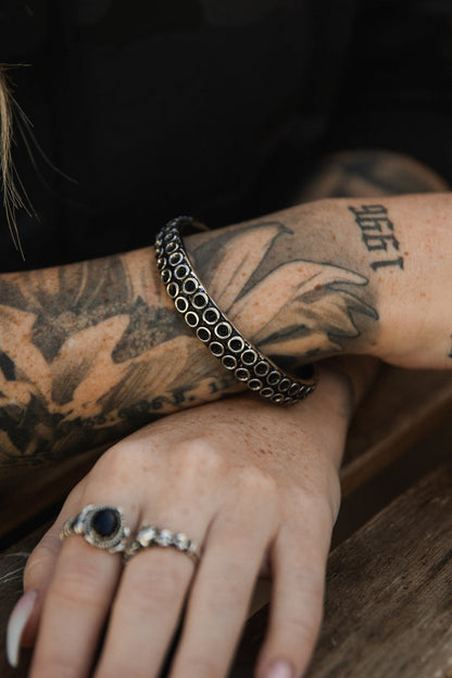 A forearm with black tattoos wears Relentless Betrayals The Kraken Bracelet. The hand, resting on wood, displays a silver ring with a dark stone and a matching band on the index finger. A blurred background enhances the intricate tattoo and jewelry details.