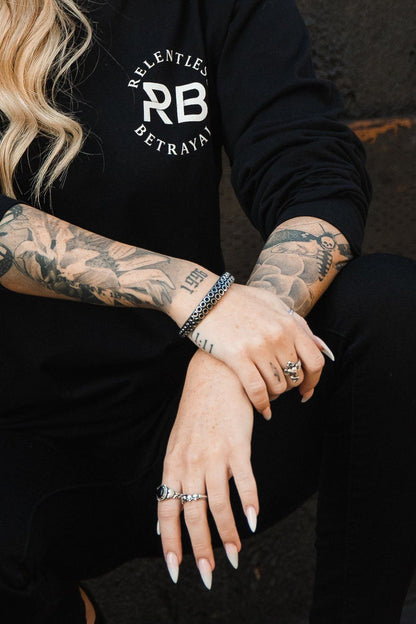 A person with long blonde hair wears a black sweatshirt featuring Relentless Betrayal RB. Their tattooed arms are crossed, showcasing rings and The Kraken Bracelet by Relentless Betrayal in hand-forged stainless steel against a dark, textured background.