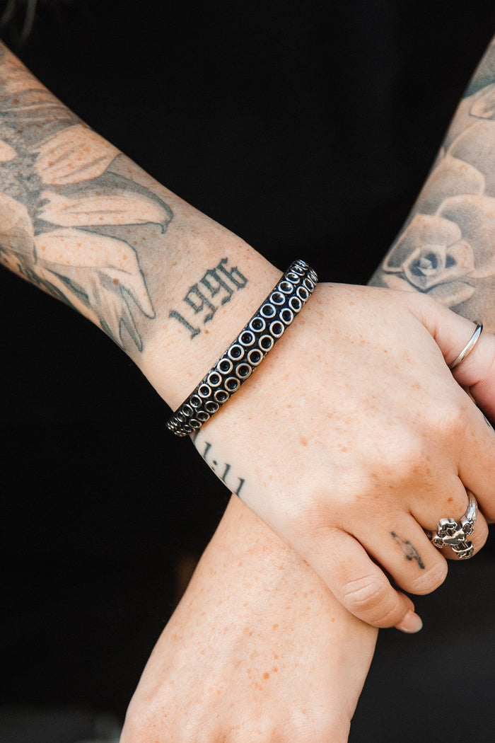 Tattooed hands with 1996 on one wrist and decorative ring tattoos, clasped against a black background. They are adorned by Relentless Betrayals The Kraken Bracelet, showcasing a circular pattern, enhancing the elegance of the scene.