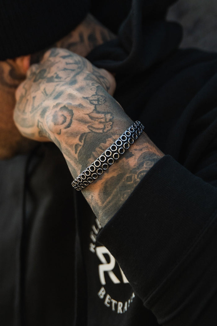 A tattooed arm with intricate designs rests near a face wearing a black beanie and hoodie. The arm sports Relentless Betrayals hand-forged stainless steel Kraken Bracelet. The hoodie, showing part of a white logo, creates an edgy and modern vibe.