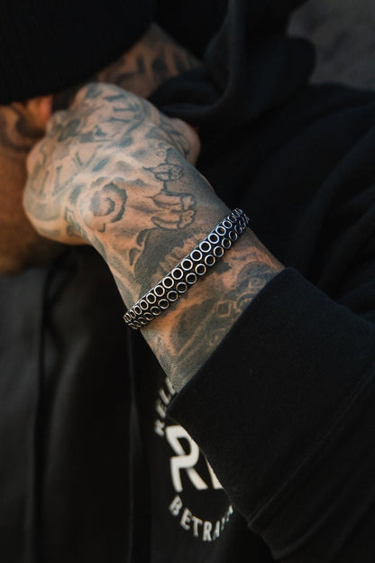 A tattooed arm with intricate designs rests near a face wearing a black beanie and hoodie. The arm sports Relentless Betrayals hand-forged stainless steel Kraken Bracelet. The hoodie, showing part of a white logo, creates an edgy and modern vibe.