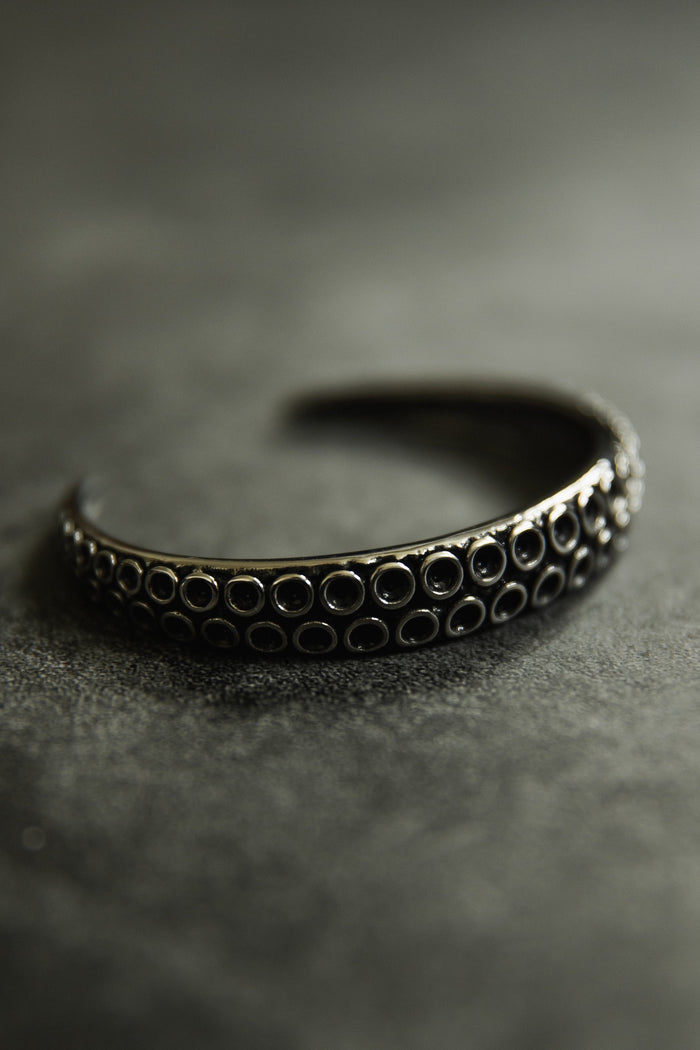The Kraken Bracelet by Relentless Betrayal showcases a hand-forged stainless steel design with a textured surface. Circular indentations adorn the partially open cuff, which rests beautifully on a smooth, dark surface.