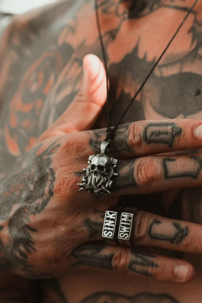 Tattooed hands grip The Kraken Necklace by Relentless Betrayal, a handcrafted pewter pendant with a skull design. The tattooed fingers feature two lettered rings, and the chain suspends the pendant against a chest inked like the mythical krakens embrace.