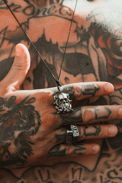 A close-up of tattooed hands and chest showcases The Kraken Necklace by Relentless Betrayal. One ring-adorned hand rests on a tattooed chest featuring skulls, flowers, and abstract designs, enhancing the intricate detail of the handcrafted pewter pendant.