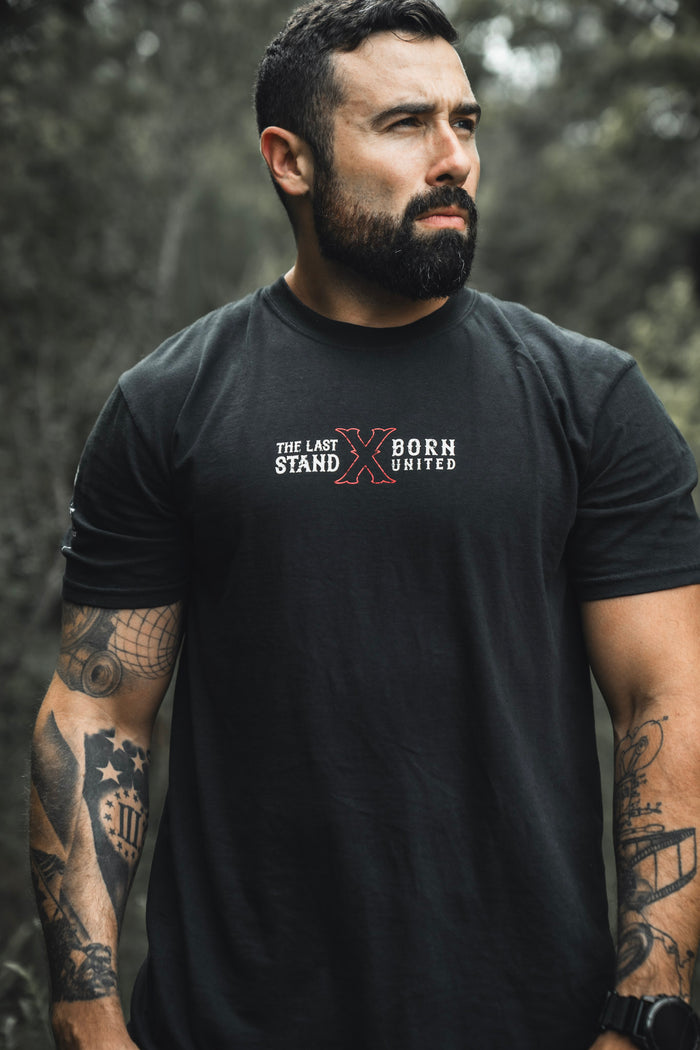 A bearded, tattooed man stands outdoors wearing The Last Stand tee from Born United Private Label, looking serious amid blurred greenery.