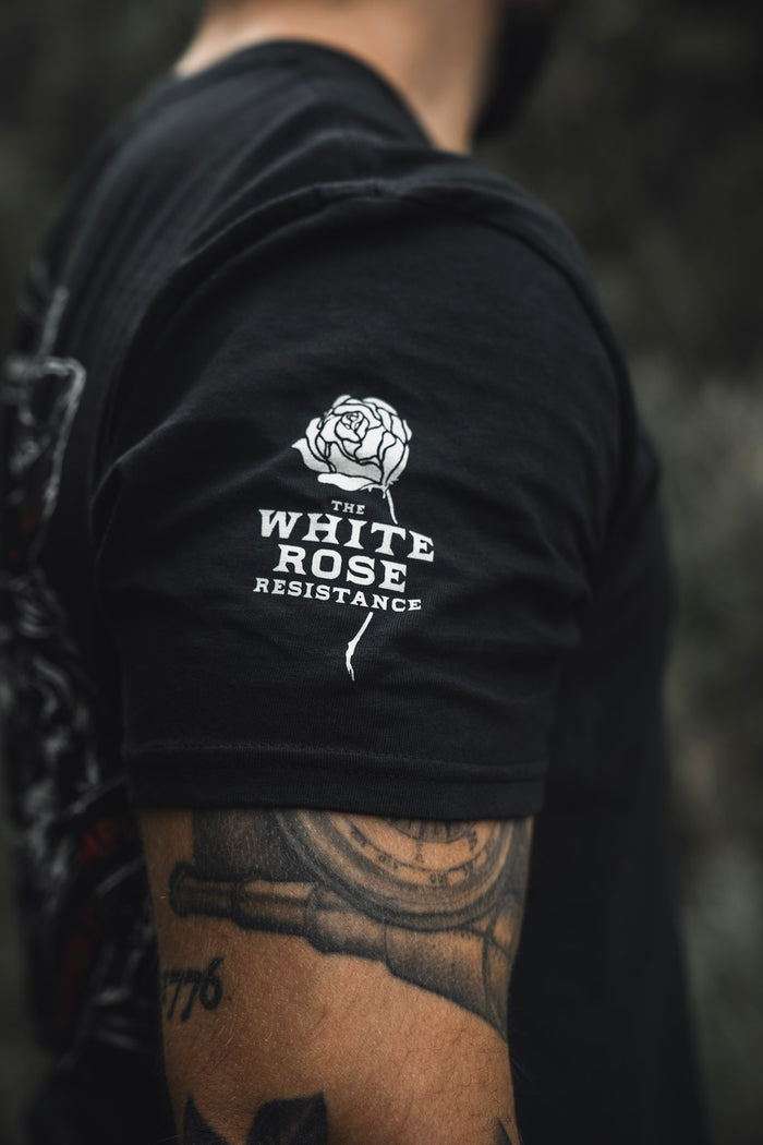 A person wears The Last Stand tee by Born United Private Label, a black shirt with a white rose graphic and THE WHITE ROSE RESISTANCE on the sleeve. Their arm tattoo stands out against the blurred outdoor background at The Last Stand Festival.