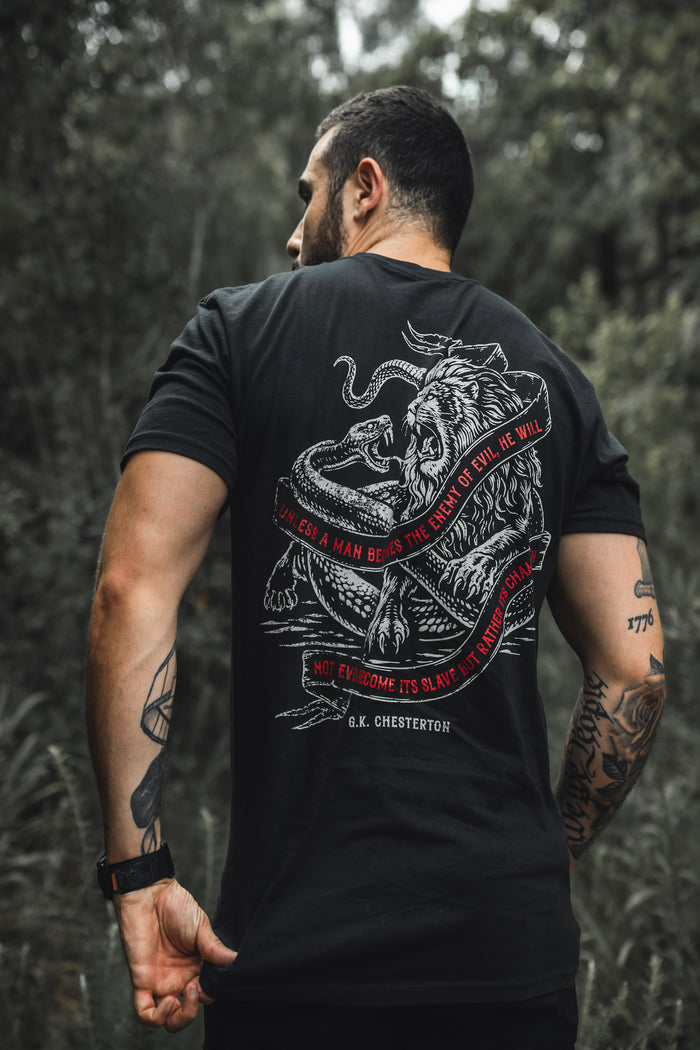 A tattooed man stands outdoors, facing away, wearing The Last Stand tee by Born United Private Label—black with a lion and snake graphic, red circular border, and G.K. Chesterton quote. Blurred trees fill the background.