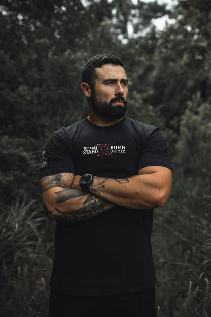 A tattooed, muscular man stands outdoors among greenery, arms crossed and looking to the side. He wears "The Last Stand" tee by Born United Private Label and a matching watch.