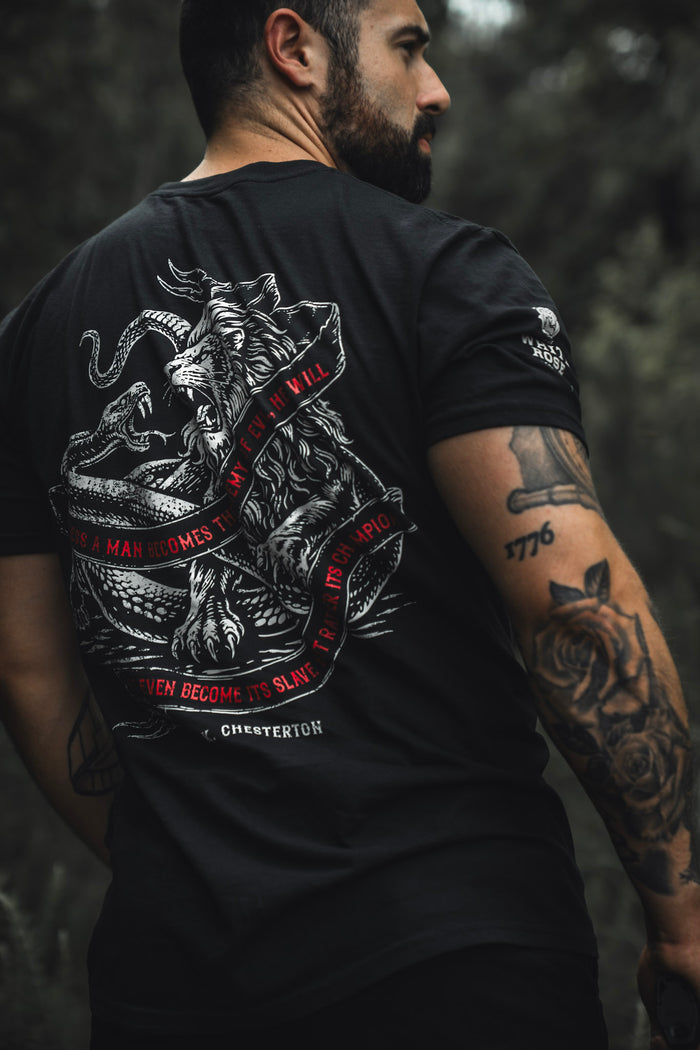 A tattooed man stands outdoors in the Born United Private Label "The Last Stand" tee, featuring a lion battling a serpent and a red-quoted banner, with a blurred green background evoking festival vibes.