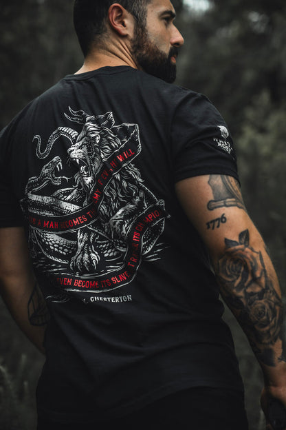 A tattooed man stands outdoors in the Born United Private Label "The Last Stand" tee, featuring a lion battling a serpent and a red-quoted banner, with a blurred green background evoking festival vibes.