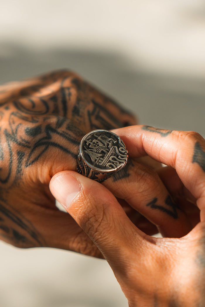 A person with tattooed hands wears a large, ornate handcrafted ring named The Rise of the Kraken by Relentless Betrayal. Engraved with a Kraken design, their fingers gently hold it against a softly blurred background.