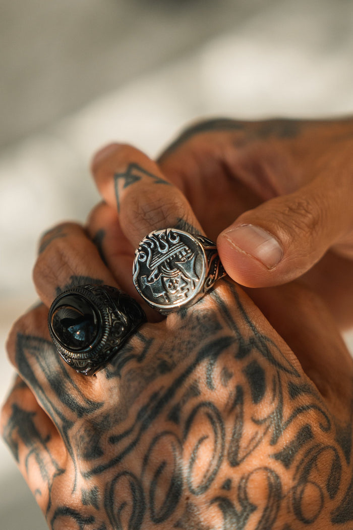 A close-up of tattooed hands features two handcrafted rings by Relentless Betrayal: one is The Rise of the Kraken Ring in stainless steel with intricate designs, and the other has a round face adorned with a dark stone.