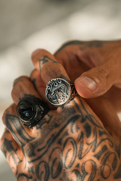 A close-up of tattooed hands features two handcrafted rings by Relentless Betrayal: one is The Rise of the Kraken Ring in stainless steel with intricate designs, and the other has a round face adorned with a dark stone.