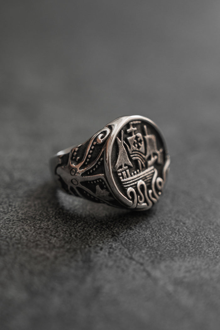 The Rise of the Kraken Ring by Relentless Betrayal, a handcrafted silver piece with detailed engravings of a sailing ship and swirling wave patterns, evocative of a mythical Kraken, rests on a dark textured surface.