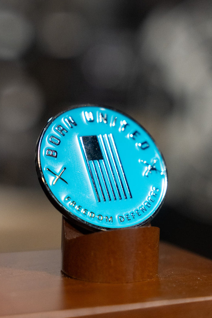 The Born United Private Label Shallows Challenge Coin features an American flag, crossed swords, and “Born United” and “Freedom Defender,” standing upright on a wooden stand—a reminder to never ask, "Bury me in the shallows.