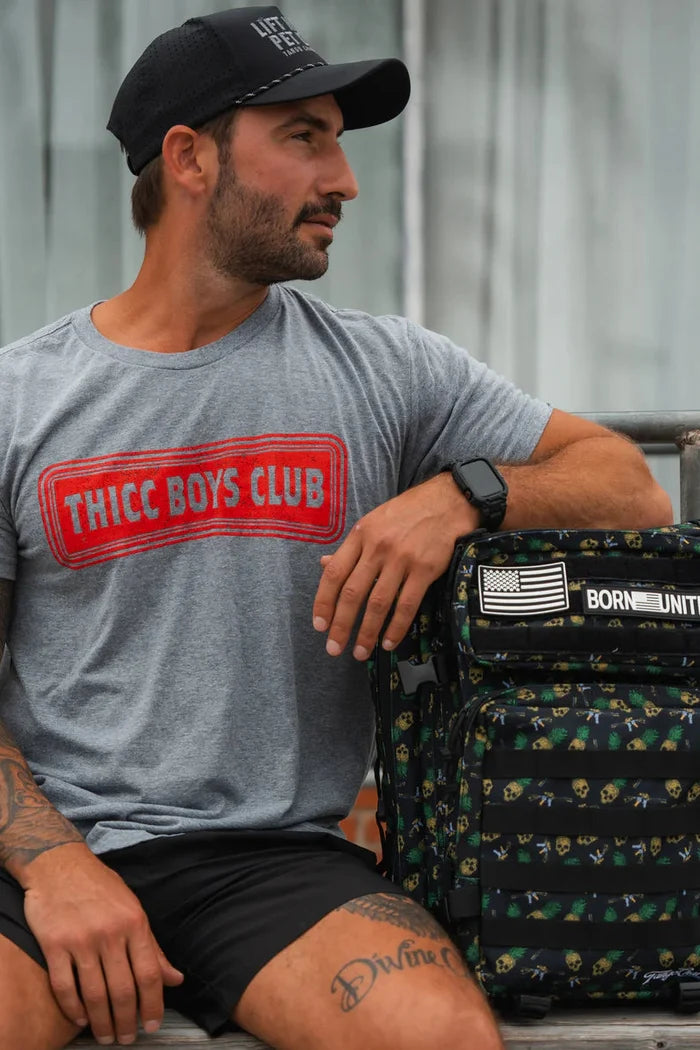 A man in a black cap and gray Tango Charlie Apparel THICC Boys Club T-Shirt sits on a bench with his arm on a black backpack featuring an American flag patch, looking left—ready to lift or just showing its Time To Eat.