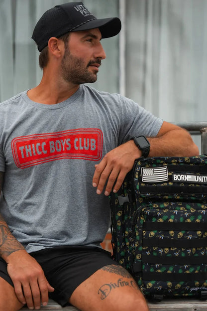 A man in a black cap and gray Tango Charlie Apparel THICC Boys Club T-Shirt sits on a bench with his arm on a black backpack featuring an American flag patch, looking left—ready to lift or just showing its Time To Eat.