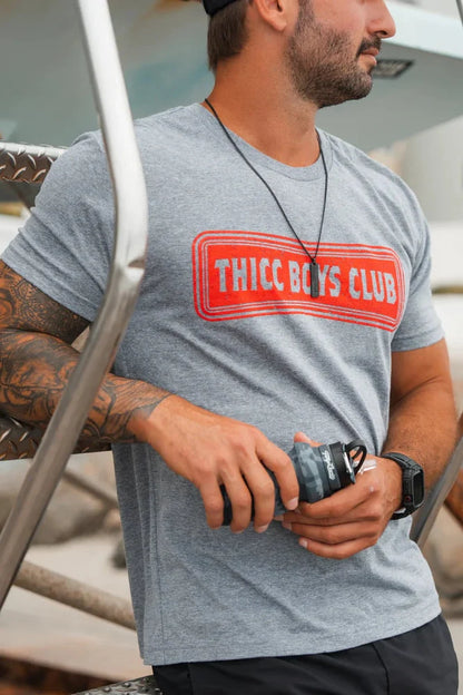 A bearded, tattooed man in a gray Tango Charlie Apparel THICC Boys Club T-Shirt and black cap leans on metal stairs with a camera lens, standing outdoors by boats—ready to lift after getting the shot.